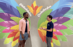 Malik and Jay outdoors in front of a colorful feathers mural