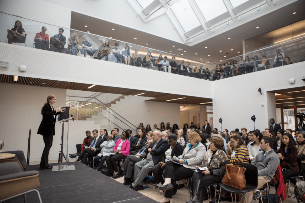 A speaker presents to a full room with people seated at ground level and on a second floor.